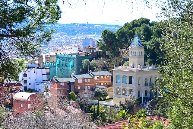 A stepped path winding down through trees and shrubs near the top of Park Güell, with the rooftops of Barcelona in the distance